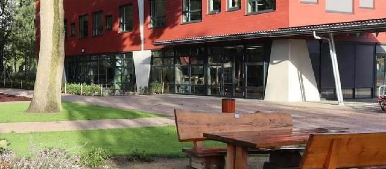 Nieuwbouw van basisschool Den Doelhof in Meijel met rood beklede gevel en grote ramen; voor het gebouw liggen houten picknickbanken op het groene schoolplein.