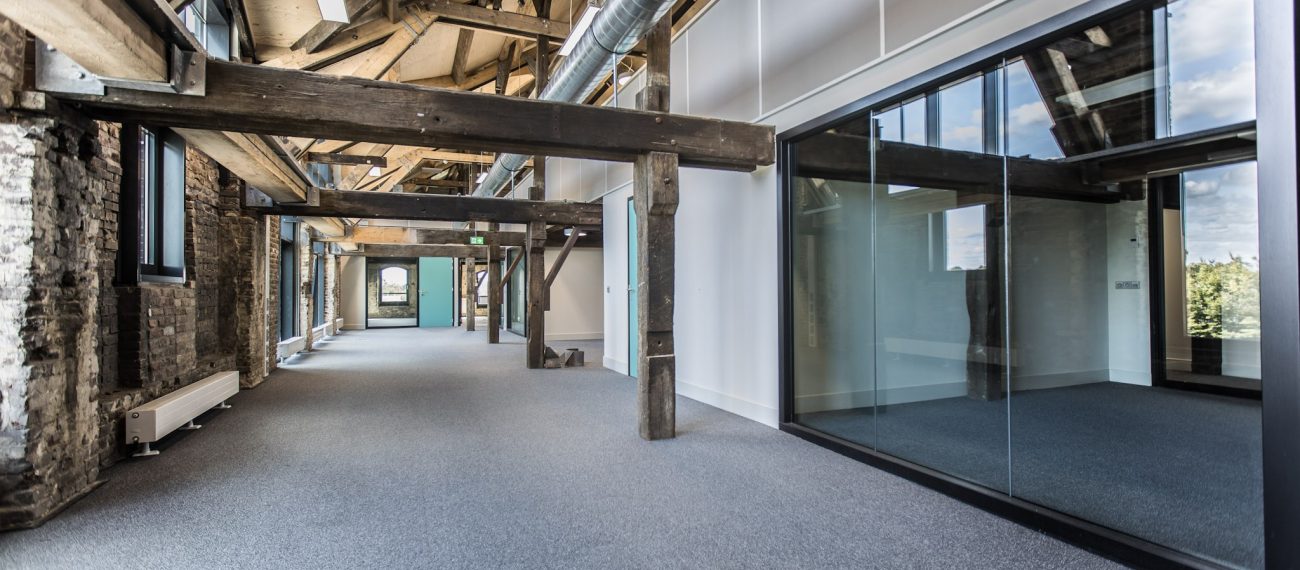 Industriële gang met zichtbare houten balken en bakstenen wanden, glazen kantoorruimtes rechts, zichtbare luchtkanalen onder het schuine dak en grijze tapijtvloer.