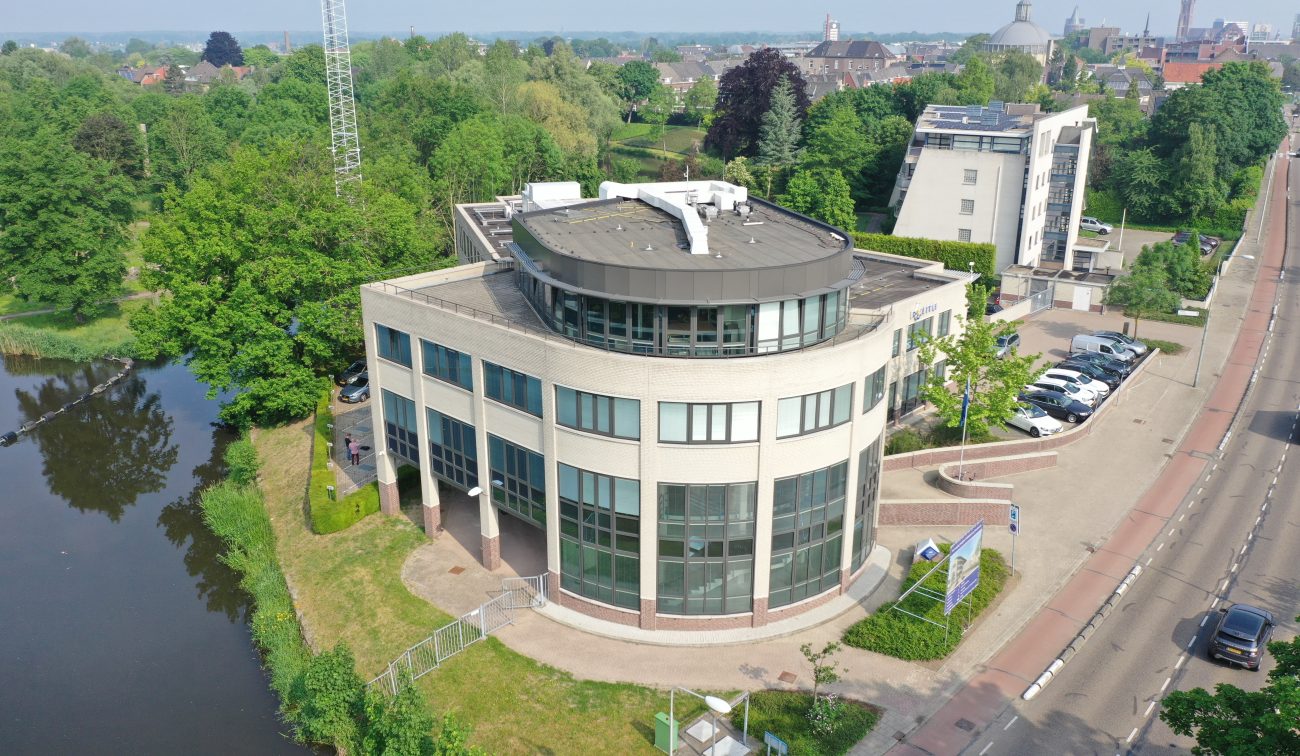 Luchtfoto van het gerenoveerde politiebureau in Roermond met ronde glazen hoekgevel, donkergrijze dakopbouw en plat dak, gelegen aan het water met parkeervakken en toegangsweg rechts.