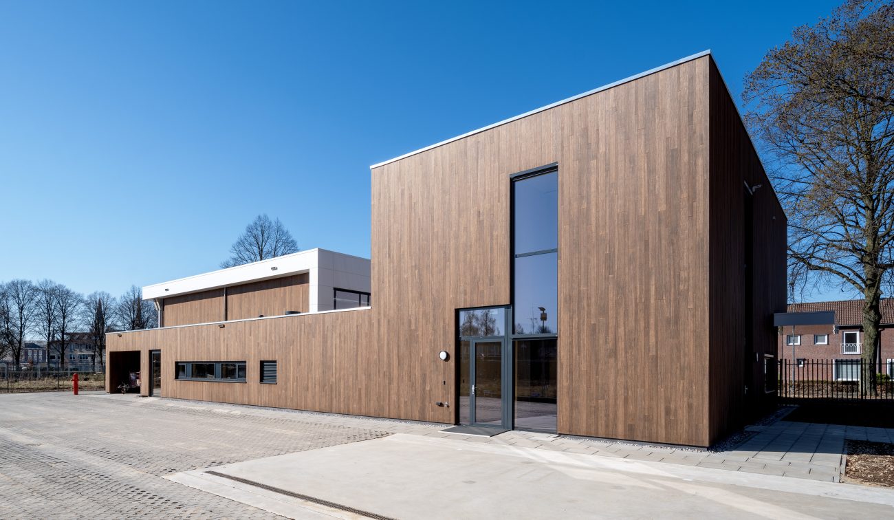 Dagopname van de nieuwe brandweerpost Sittard-Oost met houtbeklede gevel, hoge glazen entree en wit volume erachter, aan een ruim verhard voorterrein onder een strakblauwe lucht.