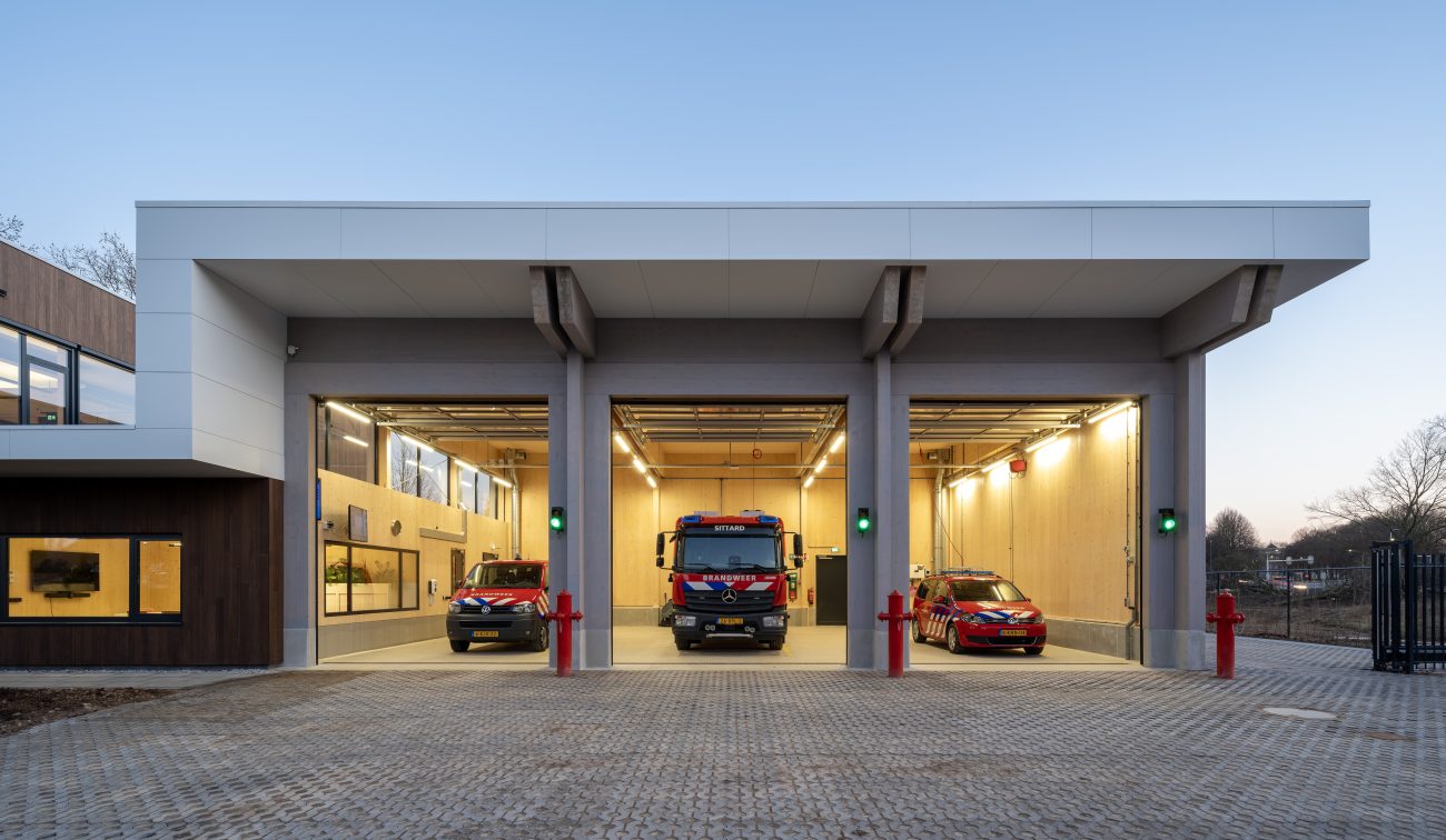 Front van de nieuwe brandweerpost Sittard-Oost in de schemering met drie open uitrijpoorten; links een brandweerbus, midden een tankautospuit en rechts een dienstauto, allemaal klaar voor uitruk op het verlichte terrein.