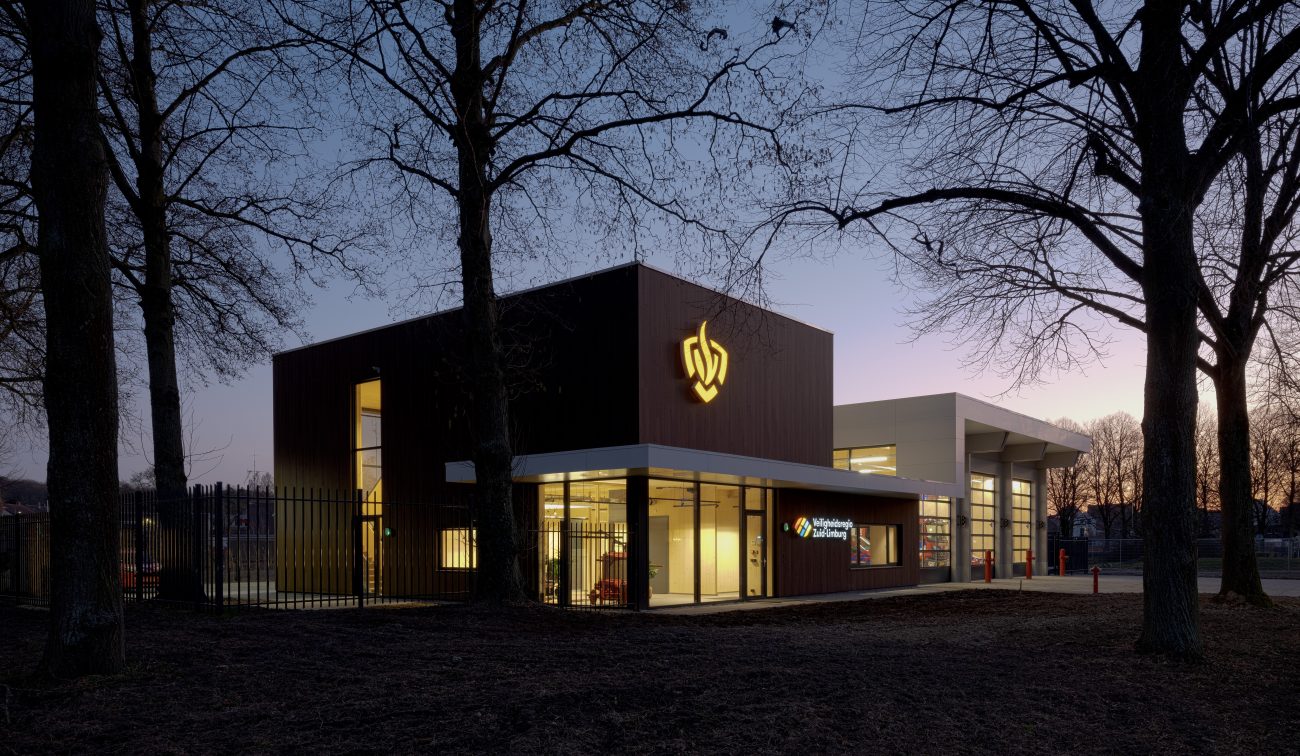 Moderne brandweerpost Sittard-Oost in de schemering met verlichte gevel en glazen entree; rechts de uitrijhal voor voertuigen, omlijst door silhouetten van bomen.
