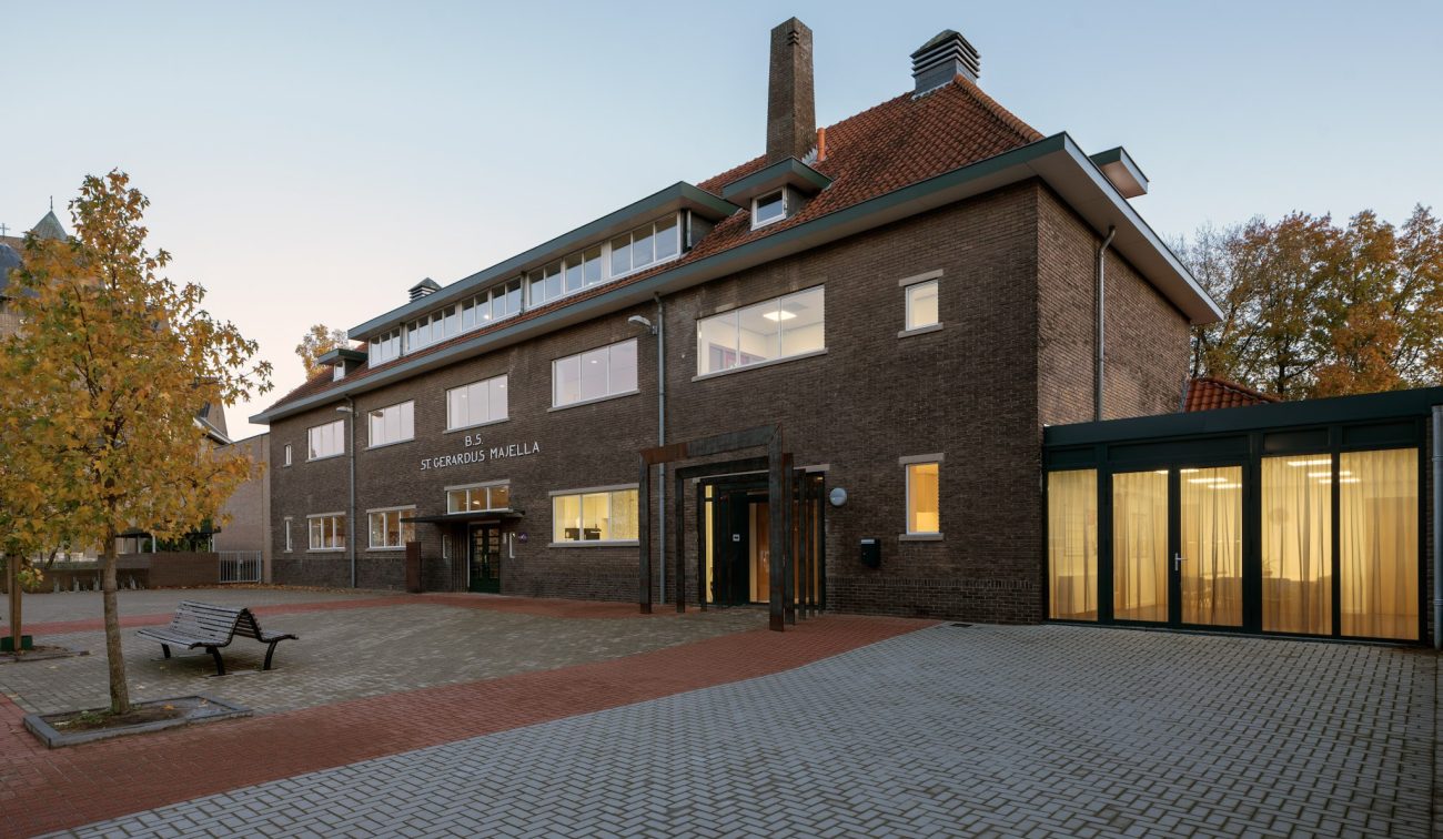 Gerestaureerd schoolgebouw St. Gerardus Majella in Heerlen met donkerbruine bakstenen gevel en rood pannendak, vernieuwde entree en moderne glazen uitbouw met gordijnen, gezien vanaf het betegelde schoolplein met boom en bankje in avondlicht.