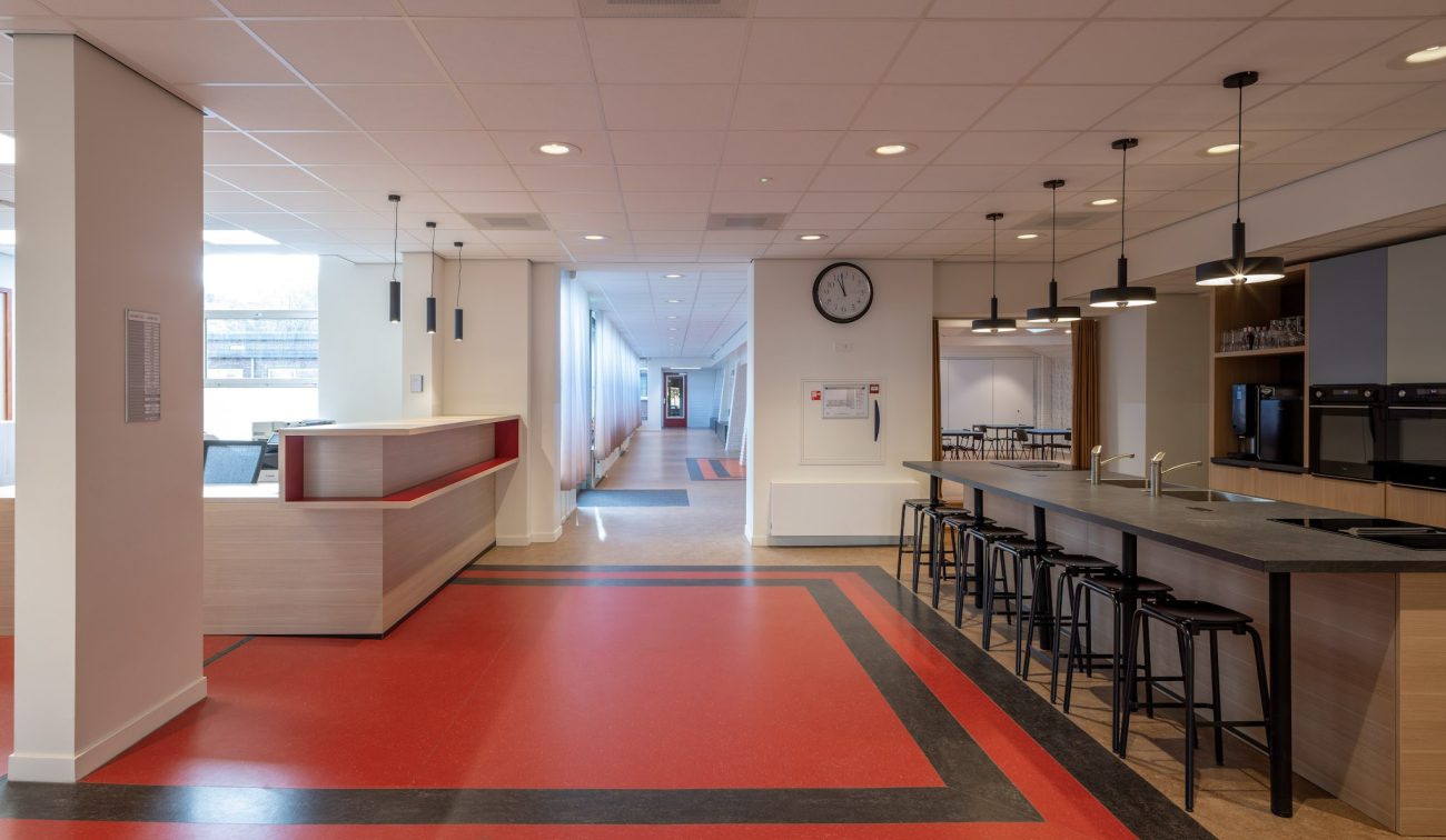Centrale hal van de Majella-school met rode en zwarte vloerbanen, ontvangstbalie links, koffiebar met hoge krukken en hanglampen rechts, klok aan de muur en lange gang met daglicht.