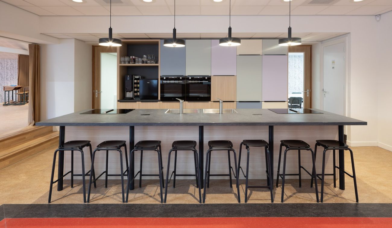 Kook/eetruimte Majella-school met groot kookeiland, acht zwarte barkrukken, vier hanglampen en pastelkleurige kastenwand met inbouwovens en koffiecorner.