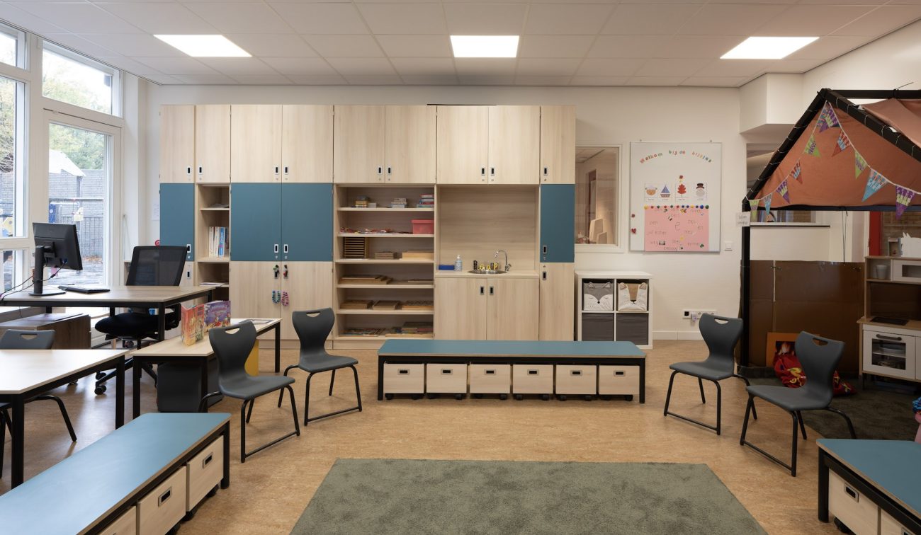 Klaslokaal Majella-school Heerlen met lichte houten kasten (blauwe accenten), lage banken met opbergbakken, kinderstoelen rond een groen vloerkleed, werkplekken en speelhoek.