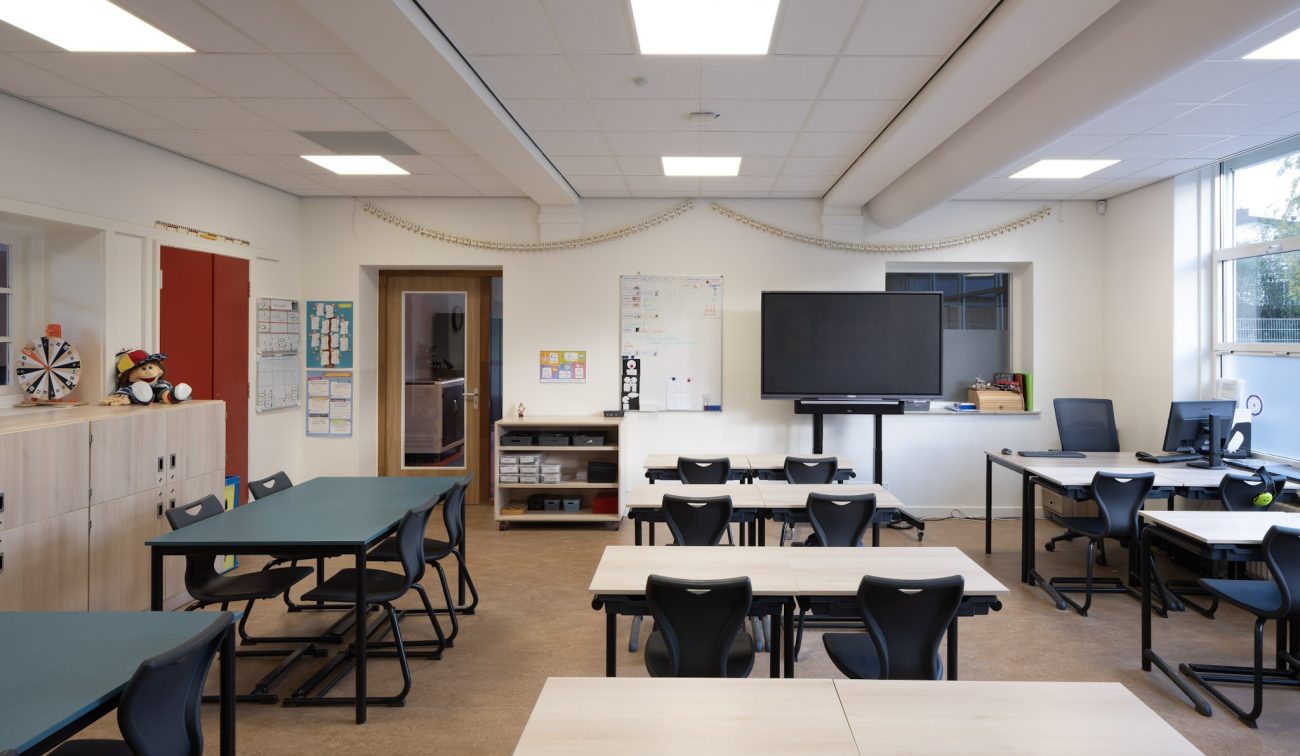 Klaslokaal Majella-school met flexibele tafels en zwarte stoelen, smartboard en whiteboard aan de muur, computers rechts en veel daglicht via grote ramen.