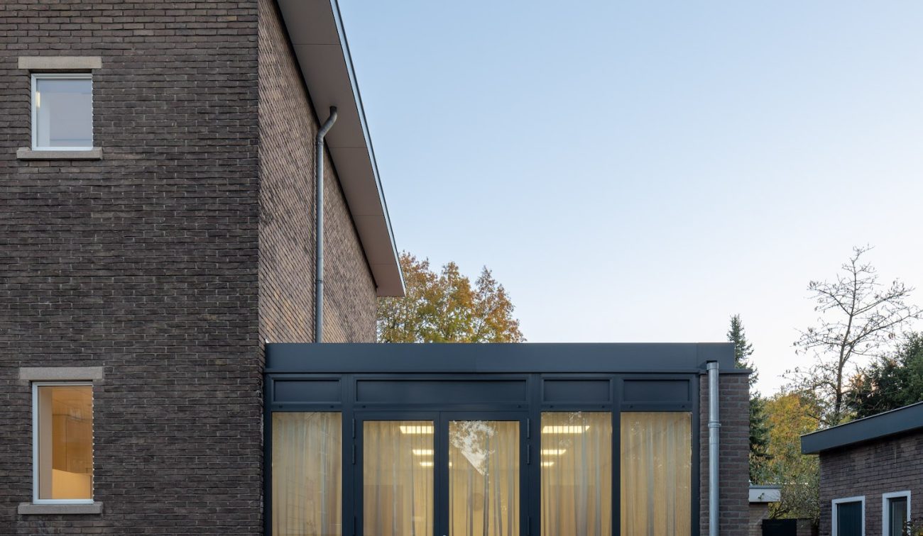 Moderne glazen aanbouw met donkere kozijnen en gele gordijnen aan de zijgevel van de gerenoveerde St. Gerardus Majella-school in Heerlen, gezien vanaf het bestraatte plein in schemerlicht.