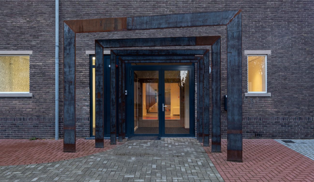 Entree Majella-school in Heerlen met cortenstalen portaalframes voor dubbele glazen deur tegen een bakstenen gevel, in schemerlicht op een patroon van klinkers.