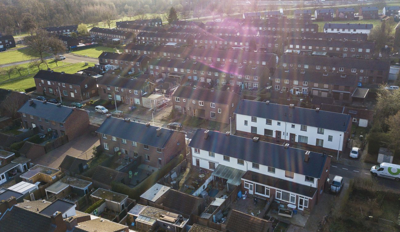 Luchtfoto van een woonwijk met rijtjeshuizen; gerenoveerde woningen met witte gevels en donkere daken steken af tussen bakstenen huizen. Zicht op achtertuinen, een straat met geparkeerde auto’s en zonnestralen in beeld.