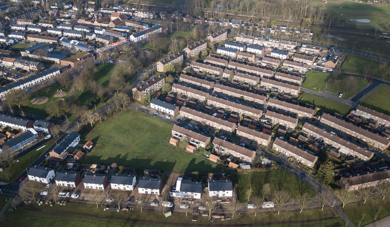 Luchtfoto van een woonwijk met lange rijen rijtjeshuizen en groene veldjes tussen de straten; renovatieblokken met lichte gevels vallen op tussen oudere baksteenwoningen.