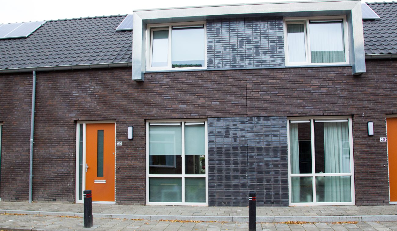 Rij moderne nieuwbouwwoningen in Zevenbergen met oranje voordeuren, strakke baksteenarchitectuur en zinken dakkapellen onder een grijs pannendak, in een rustige woonstraat zonder verkeer.
