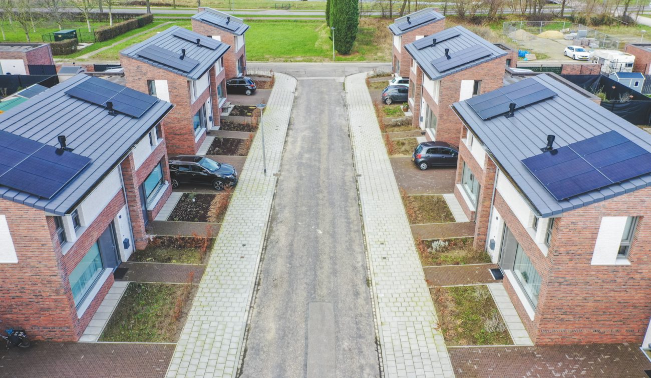 Luchtfoto van een smalle straat tussen nieuwe rijwoningen in Nazareth (Maastricht); bakstenen gevels met grote ramen, daken vol zonnepanelen, kleine voortuinen en geparkeerde auto’s langs het trottoir.