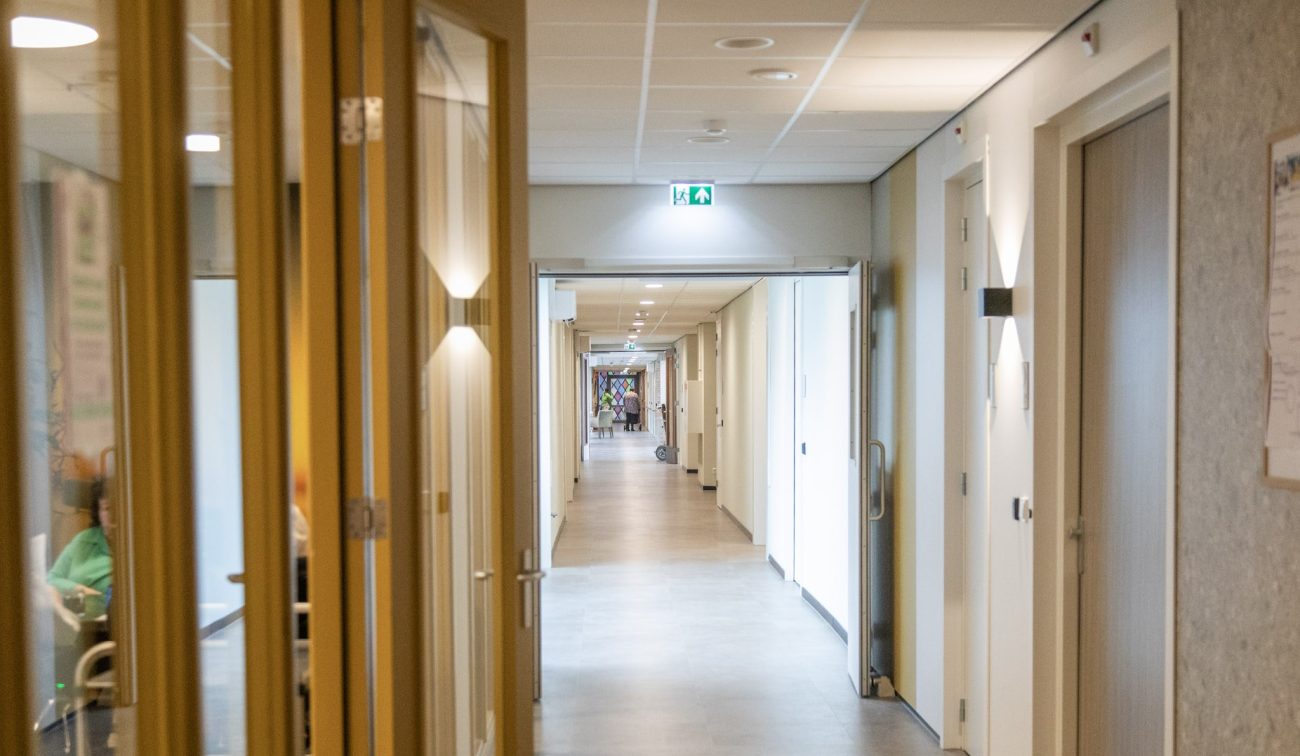 Lange, lichte zorggang met systeemplafond, nooduitgang-pictogram en deuren aan beide zijden; links een glaspui met open deuren en zicht op wachtende personen.