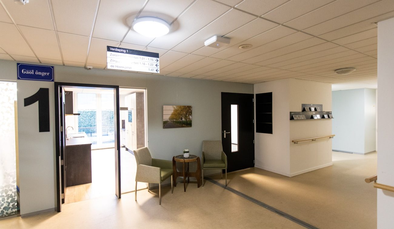 Zorghuis-gang met systeemplafond en ronde plafondlamp; grote ‘1’ bij een open deuropening naar een keuken, twee fauteuils met bijzettafel tegen een grijze wand, zwarte deur, wegwijzerbord aan het plafond en handrail langs de muur.