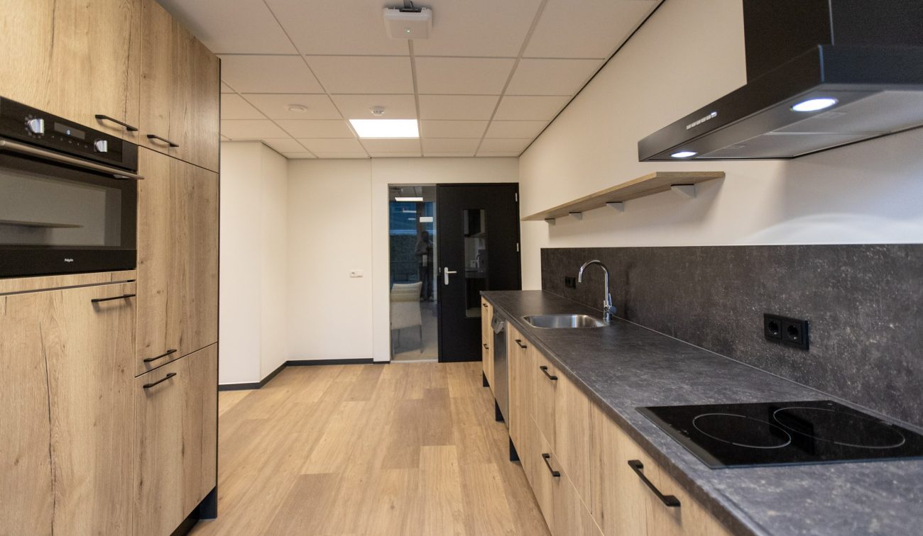 Moderne keuken met houtlook kasten, donker composiet werkblad met inductiekookplaat en rvs-spoelbak, zwevende plank aan witte wand, systeemplafond en zwarte deur naar de gang