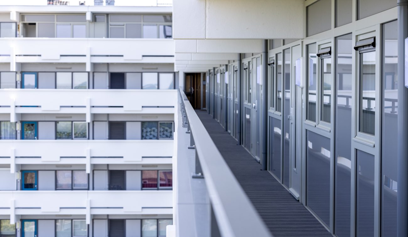 Perspectief langs een galerij met grijze voordeuren en glazen balustrade; aan de overzijde is de witte gevel met ramen en balkons van de tegenoverliggende vleugel te zien