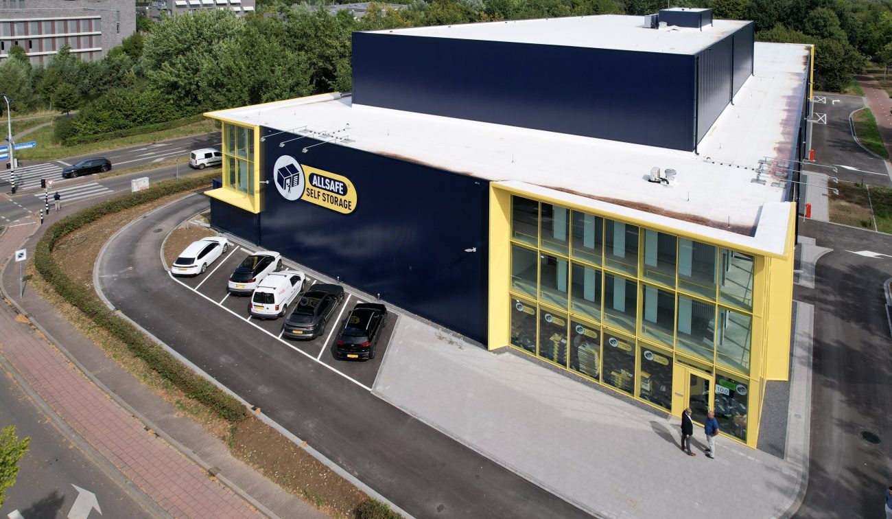 Luchtfoto van de nieuwbouw van Allsafe Self Storage in Maastricht: donkerblauw volume met felgele kaders rond de glazen hoekgevel, witte dakrand en parkeervakken met auto’s langs de voorzijde; twee personen lopen richting de entree, omringd door wegen en groen.