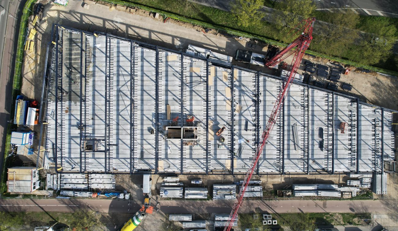 Verticale dronefoto van een bouwplaats met een trapeziumvormig gebouw in aanbouw: raster van stalen/ prefab betonnen liggers, rood kraangiek diagonaal over het beeld, stapels prefab elementen langs de randen en enkele bouwvakkers zichtbaar op de vloer.