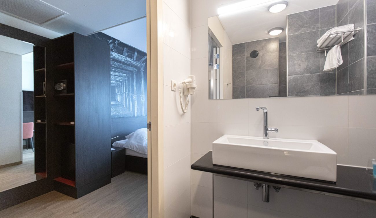 Hotelbadkamer Geleen – moderne wastafel, grijze tegelwand en zicht op slaapkamer met kast en spiegel