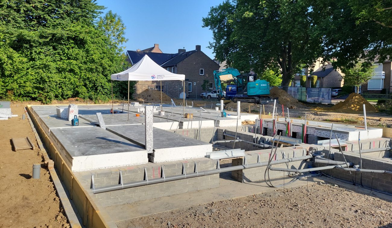 Bouwplaats Margraten: funderingsbalken met kanaalplaten, graafmachine op achtergrond.