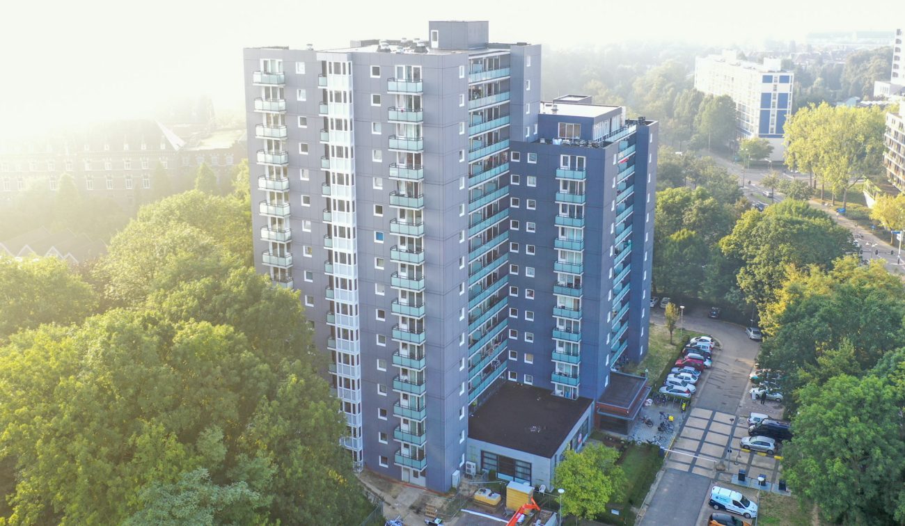 Luchtfoto van de gerenoveerde Aambosveldflat in Heerlen: twee gekoppelde grijze woontorens met glazen balkons, omringd door bomen, met parkeervakken en zacht ochtendlicht.