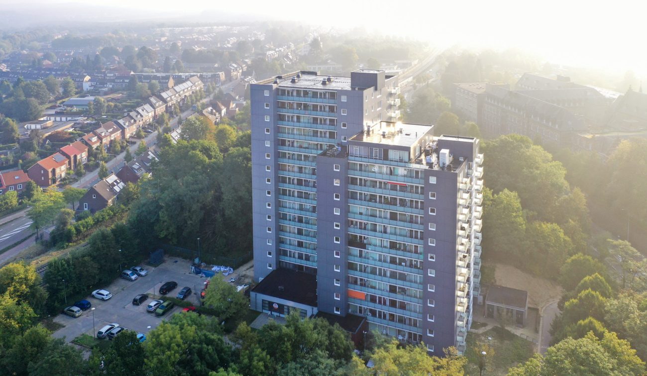 Schuine luchtfoto van de Aambosveldflat in Heerlen: twee grijze woontorens met glazen balkons, omringd door bomen, parkeerplaats vooraan en lichte ochtendnevel.