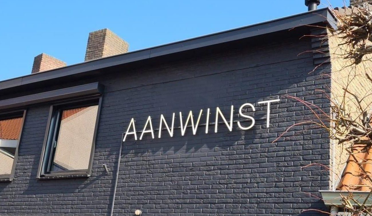 Gevel van eetcafé De Aanwinst met zwart geschilderde baksteen en grote verlichte letters “AANWINST”, onder een helderblauwe lucht.