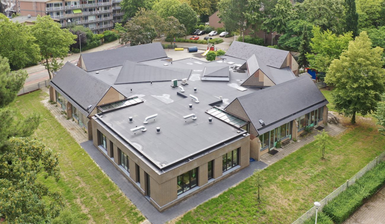 Luchtfoto van basisschool De Tovercirkel in Heerlen: gerenoveerd en uitgebreid gebouw met centrale plat dakzone met installaties en lichtstraten, omringd door paviljoens met schuine daken en grote ramen, tussen groen en speelplein.