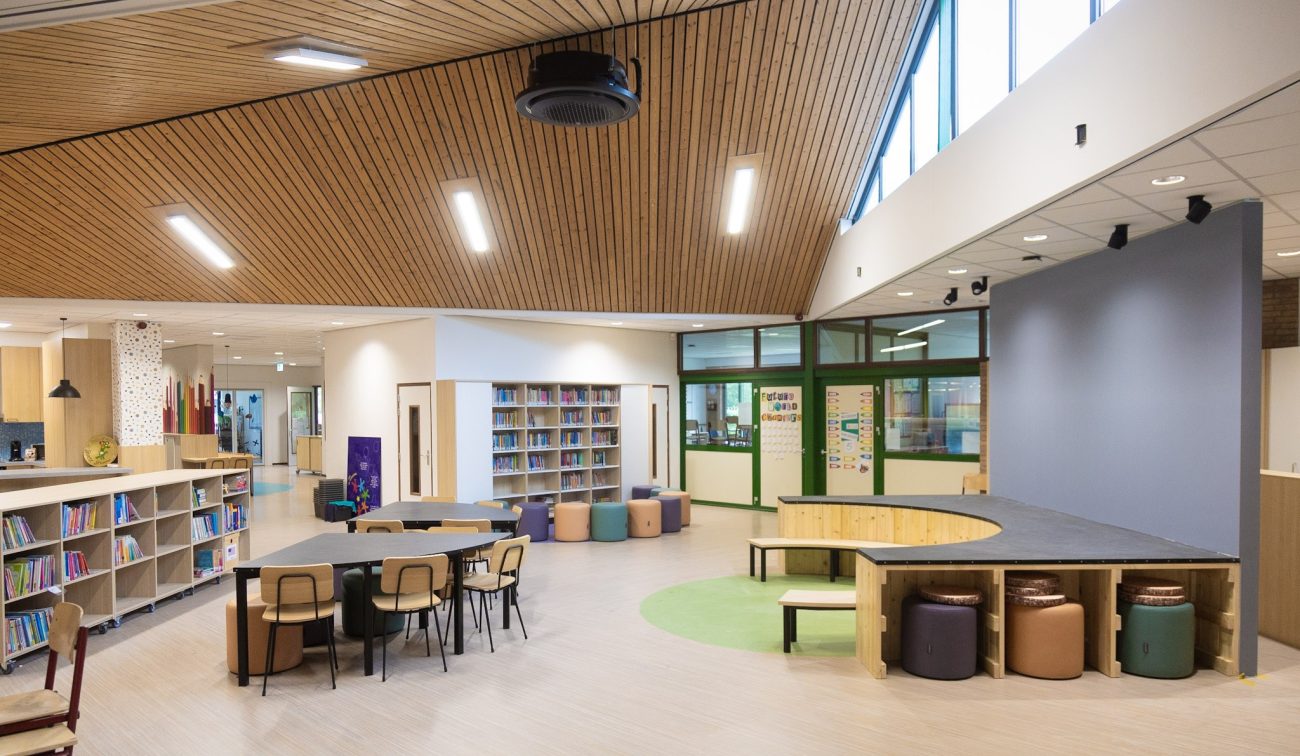 Open leerplein met houten plafond, boekenkasten en werktafels; rechts een halfrond podium met zitpoefs en groene accenten bij grote ramen.