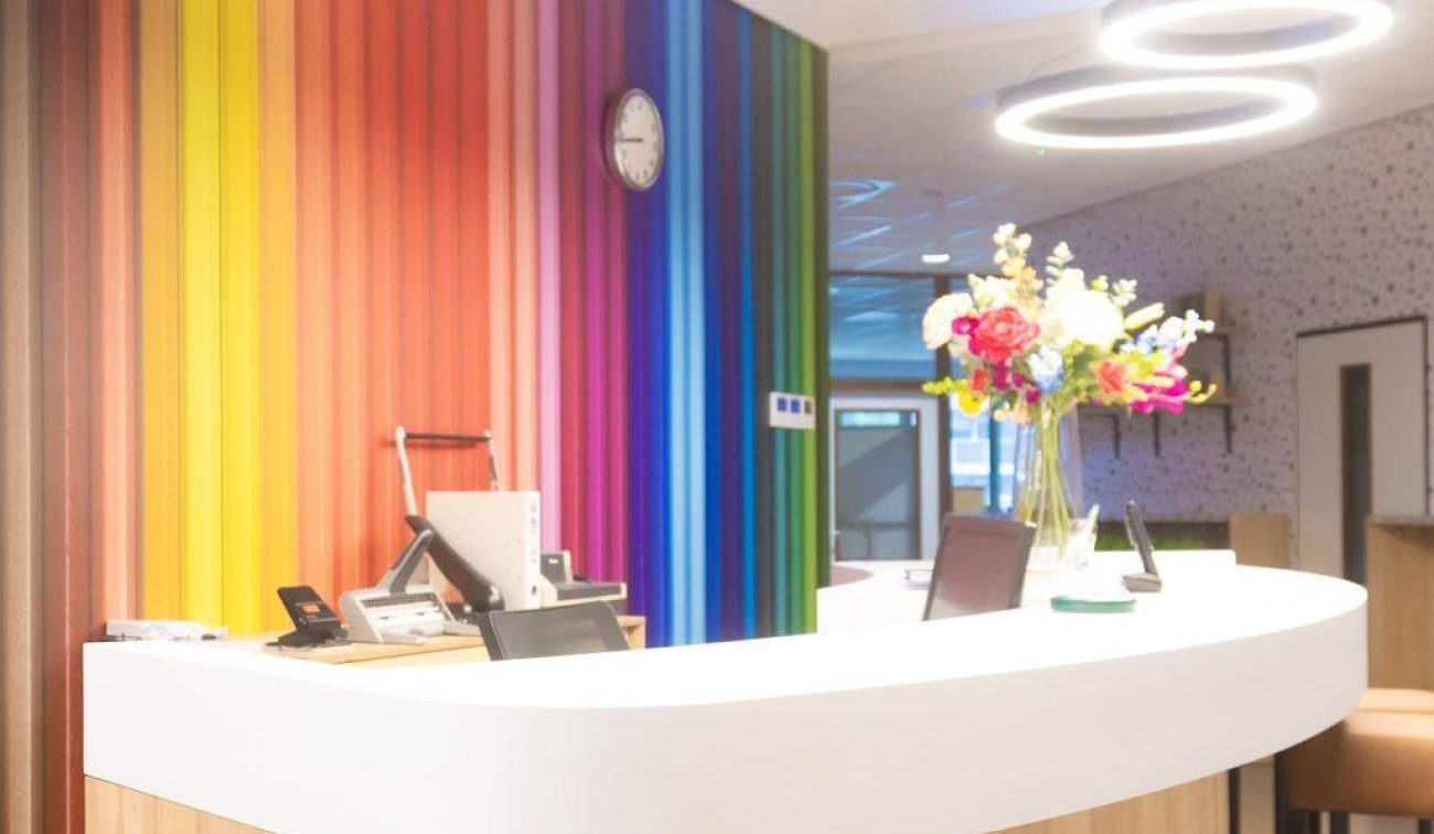 Receptiebalie met witte, afgeronde top en houten front; op de achtergrond een regenboogkleurige wand met klok en ronde ringlampen, vaas met bloemen op de balie.