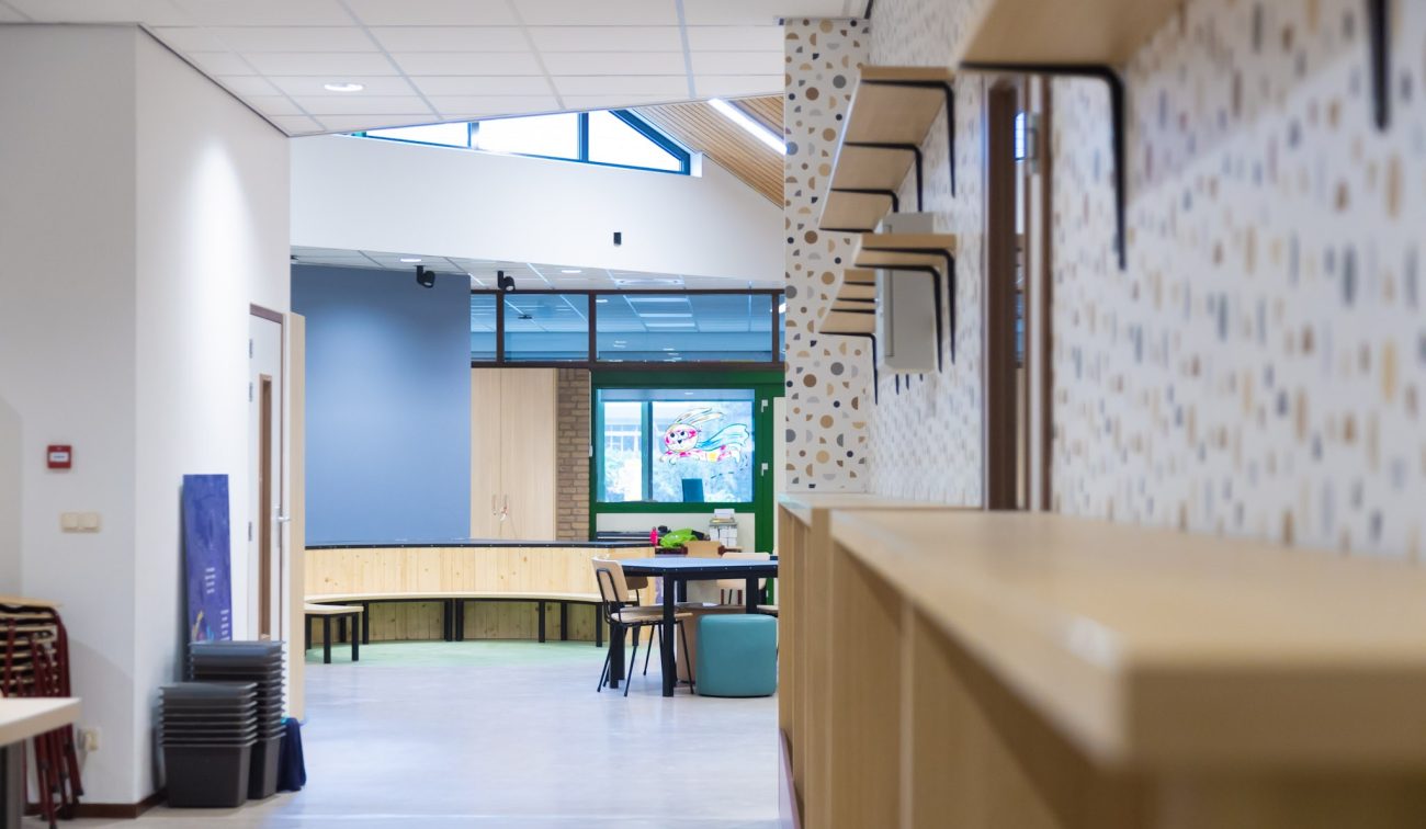 Lichte schoolgang met houten kasten en speelse wandplankjes; zicht op open leerplein met ronde tafel, krukken en grote ramen onder een schuin dak met daklicht.