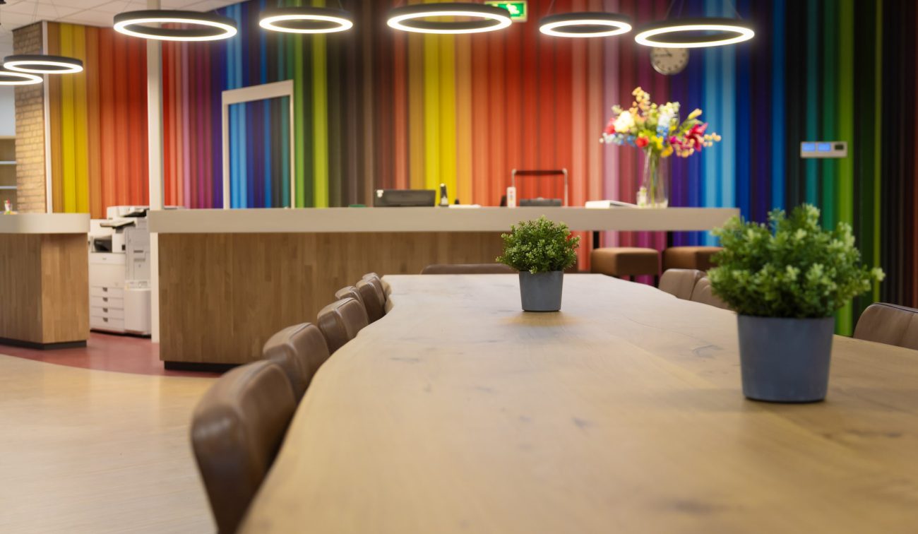 Interieur basisschool—ontvangstbalie met kleurrijke potloden-wand, ronde ringlampen en een lange houten tafel met plantjes.