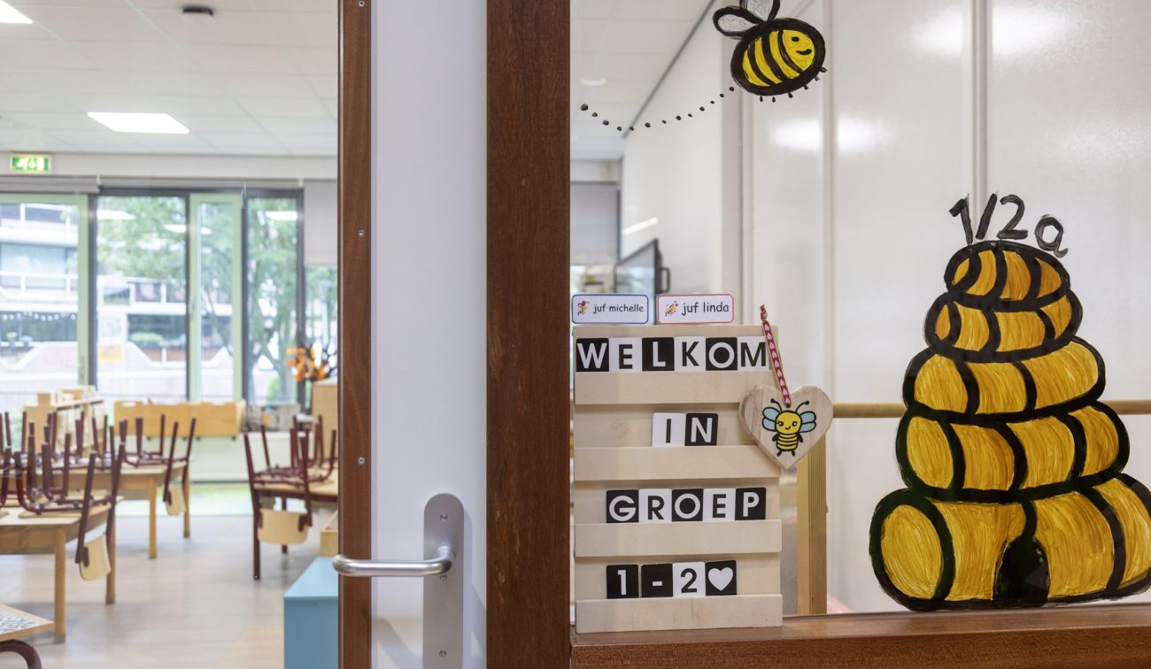 Deurkijkje basisschool—raam met tekening van een bijenkorf en bijtjes en een bord “Welkom in groep 1-2”; daarachter een klaslokaal met opgestapelde stoelen en grote ramen.