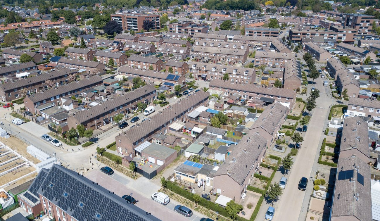 Luchtfoto van een dichtbebouwde woonwijk met lange rijen rijtjeshuizen, smalle straten met geparkeerde auto’s, achtertuinen en enkele daken met zonnepanelen; links is een bouw- of renovatieplaats zichtbaar.