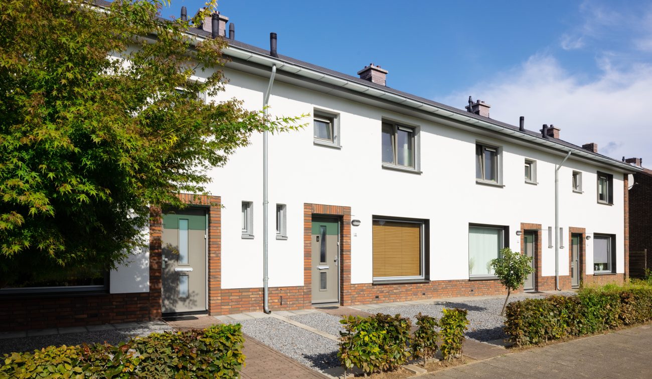 Gerenoveerde rijtjeswoningen met witte gevels en baksteenaccenten, grijze deuren en kozijnen. Voortuinen met grind en lage hagen; boom links in beeld op een zonnige dag.