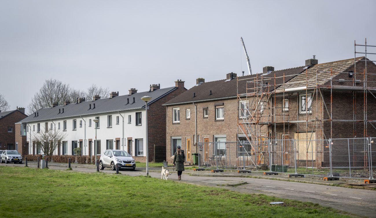 ij woningen in Maastricht; links gerenoveerde witte gevels, rechts bakstenen huizen in steigers. Een persoon met hond loopt langs en er staat een auto geparkeerd.