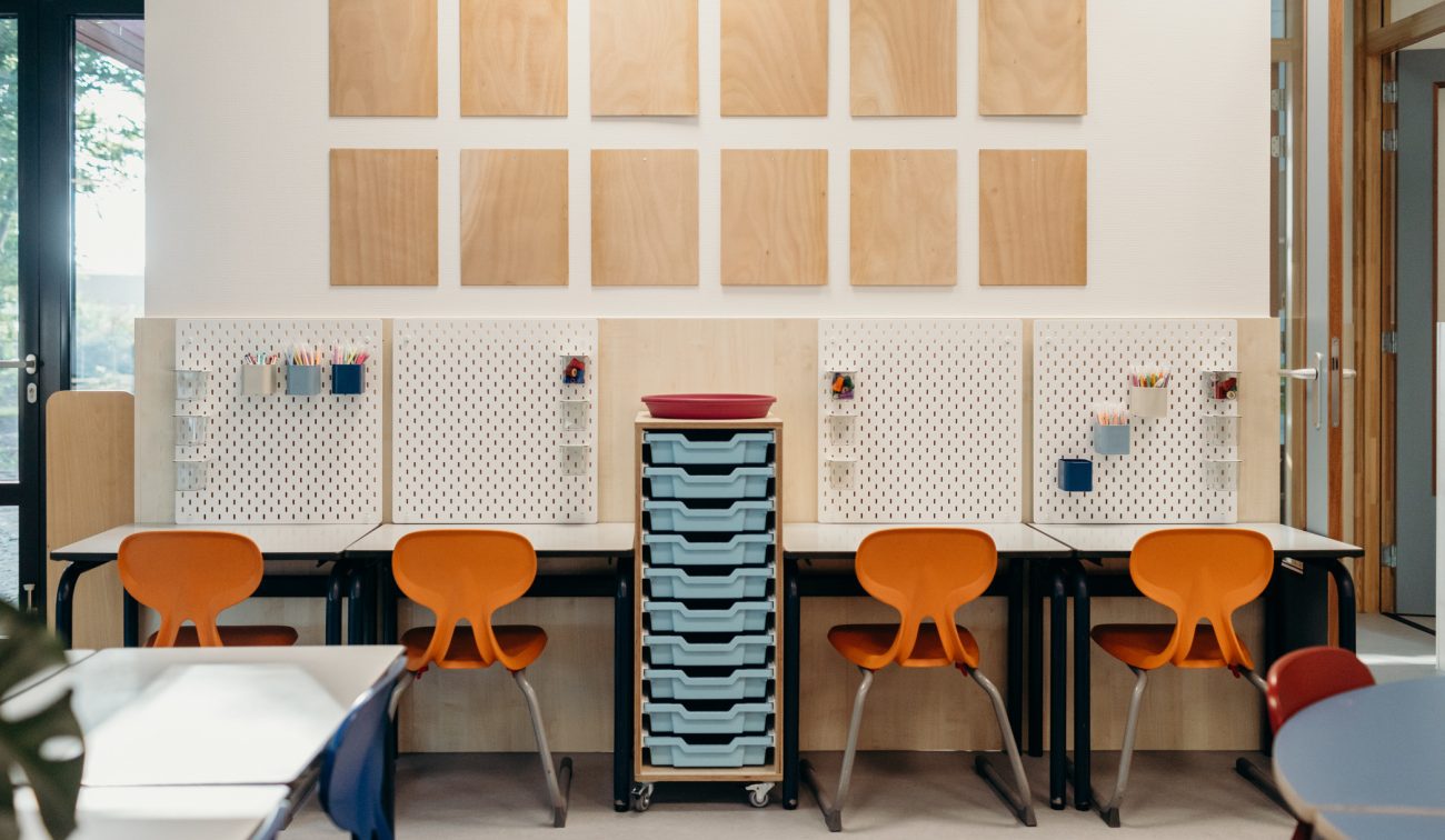 Interieur klaslokaal met vier oranje stoelen aan werkplekken met witte perforatieborden; in het midden een verrijdbare kast met lichtblauwe laden en aan de wand houten vierkante panelen.