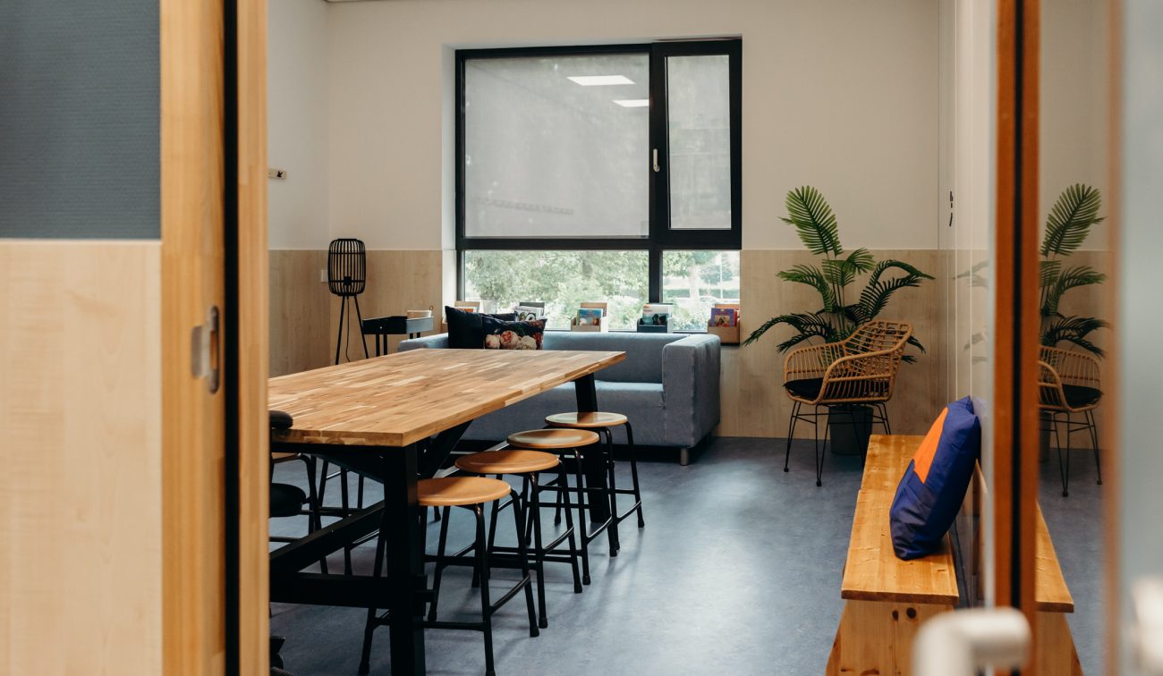 Gezellige schoolruimte met lange houten tafel en krukken, blauwe bank bij een raam met rolgordijn, twee rotan stoelen met grote kamerplanten en een houten bank met kussen op de voorgrond.