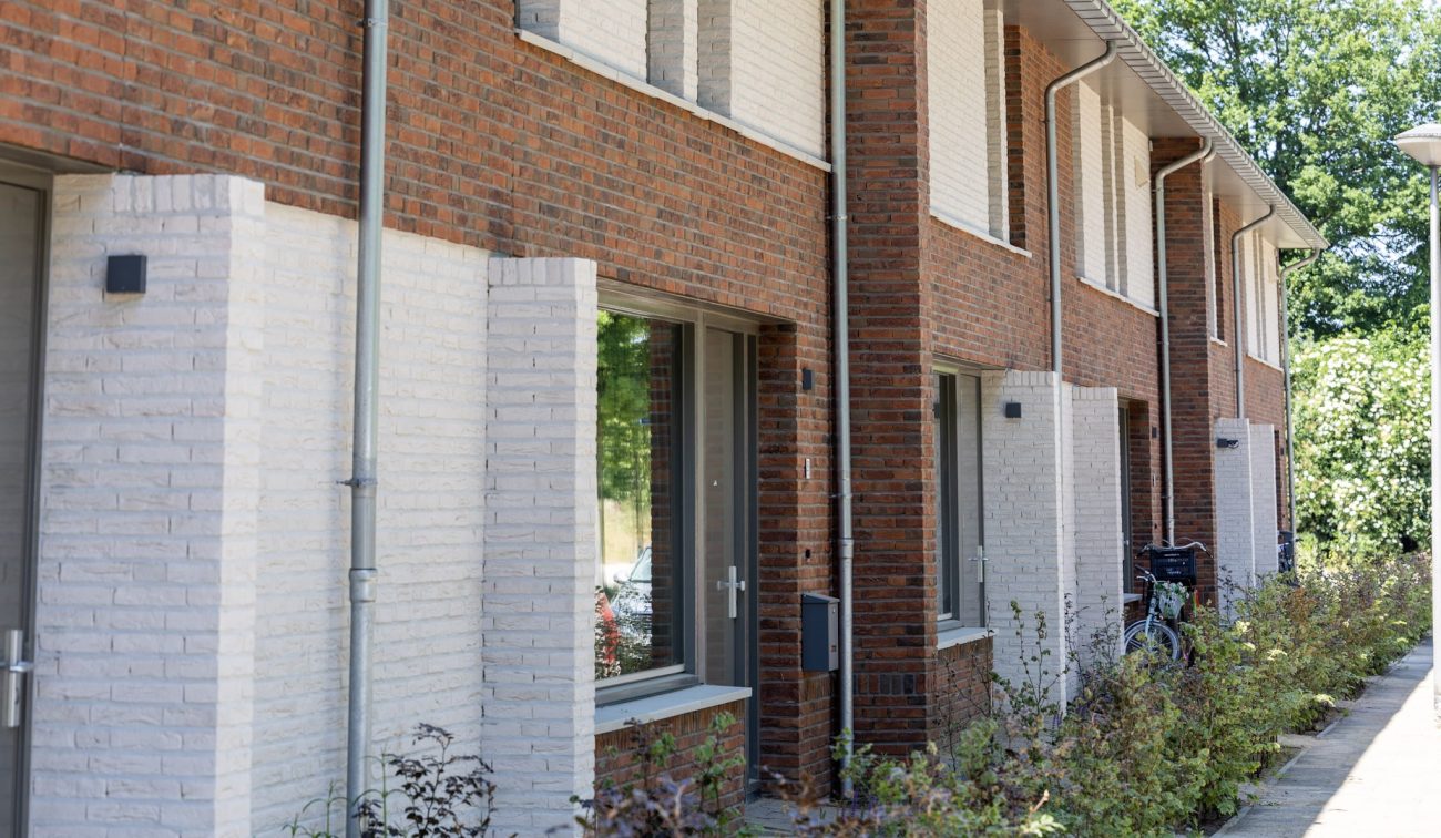 Close-up van een rij nieuwbouwwoningen met rode en wit geschilderde baksteen, grote ramen en regenpijpen; kleine voortuinen met jonge beplanting en een fiets bij een entree.