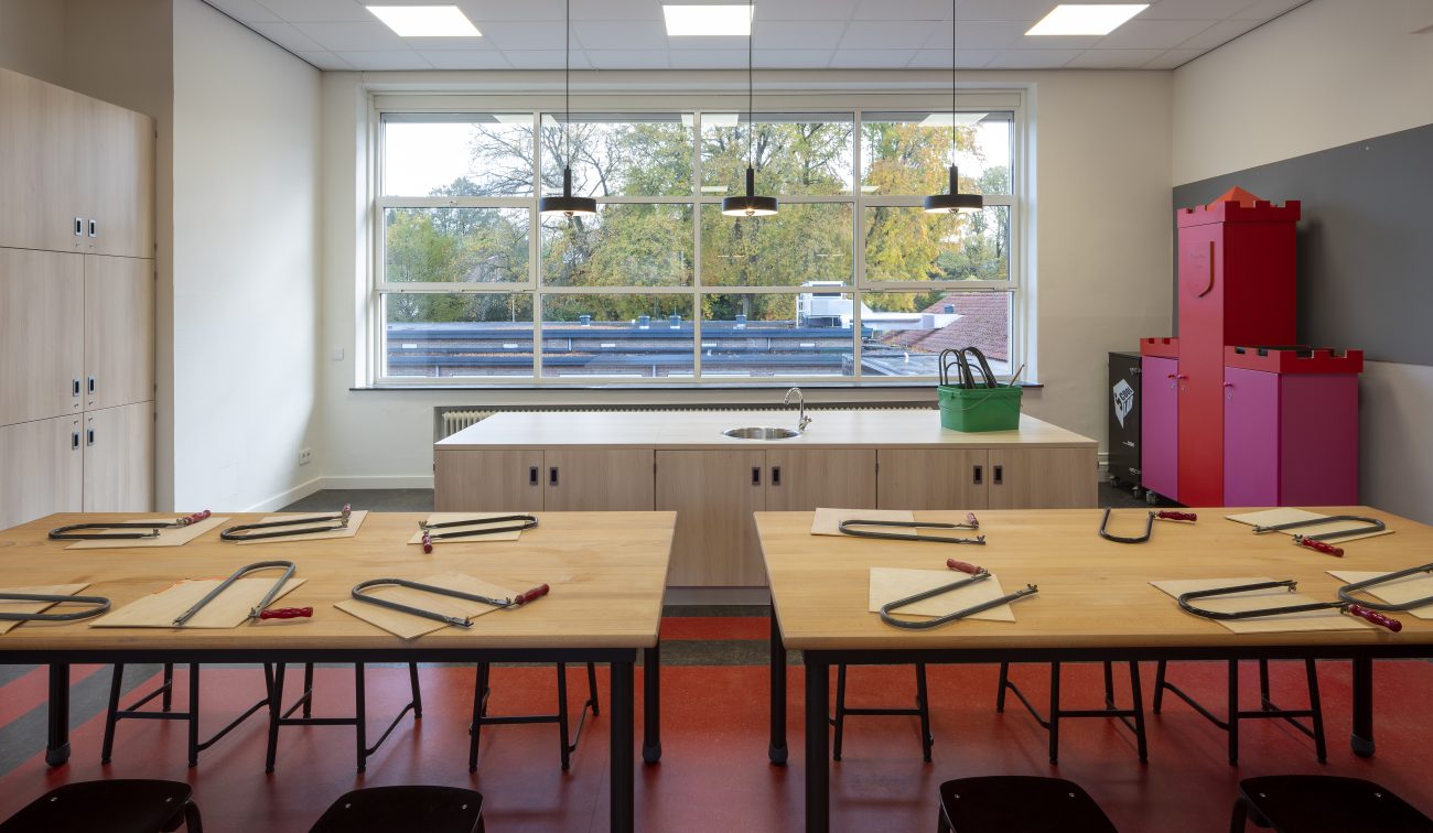 Technieklokaal Majella-school met grote werktafels vol figuurzagen, rood-zwarte vloer, centrale kast met gootsteen, grote ramen en kleurrijke opbergkasten.