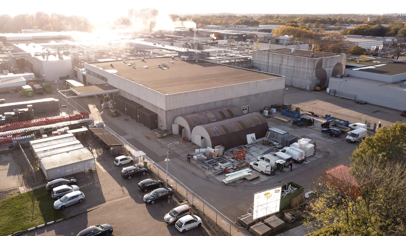 Bouwterrein bij de Mora-fabriek in Maastricht, met tijdelijke boogtenten, stapels bouwmaterialen, busjes en heftrucks naast de grote fabriekshal bij ondergaande zon.