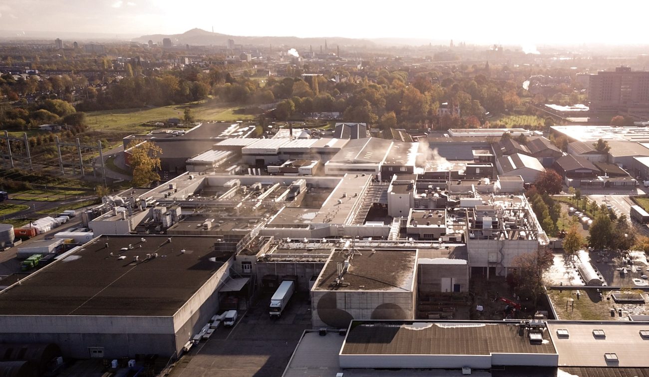 Luchtfoto van de uitgebreide en gerenoveerde Mora-fabriek in Maastricht, met daken vol installaties en stoompluimen, laadtrucks op het terrein en herfstkleurige bomen rondom.