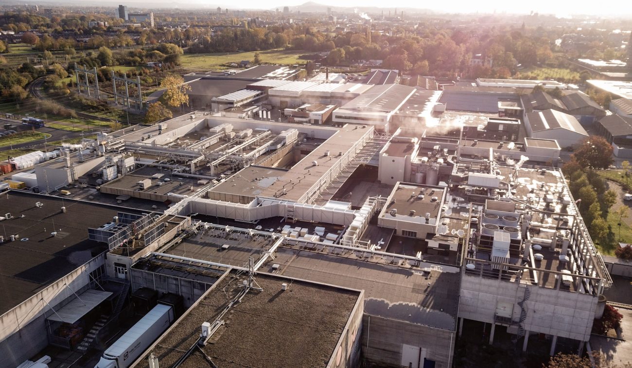 Luchtfoto van de uitgebreid en gerenoveerd ogende Mora-fabriek in Maastricht, met een uitgestrekt daklandschap vol installaties, leidingen en stoompluimen, omringd door groen en bedrijfsgebouwen.