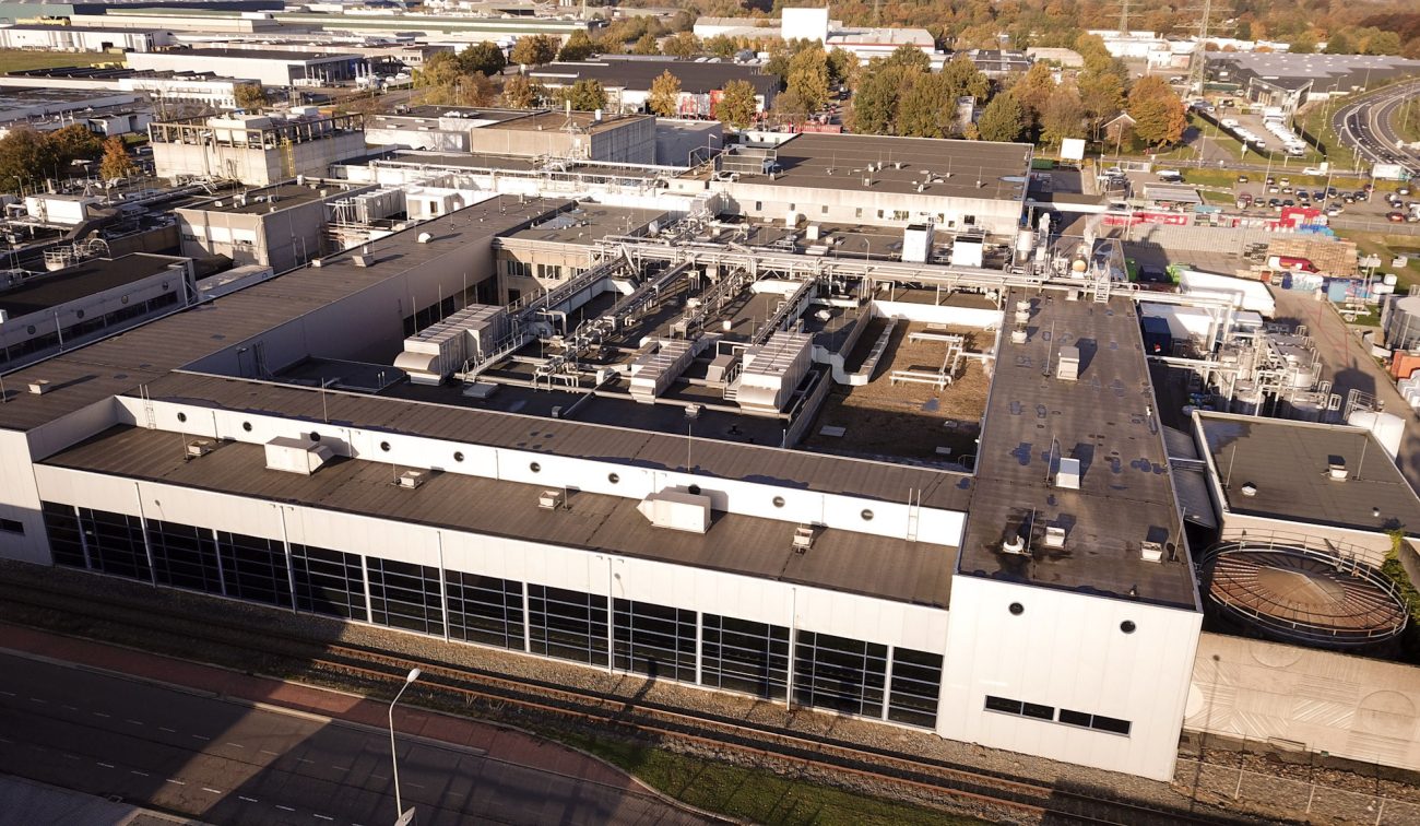 Afschuinende close-up luchtfoto van de gerenoveerde Mora-fabriek in Maastricht, met daken vol HVAC-installaties, leidingen en schachten boven een witte gevel met lange bandramen.