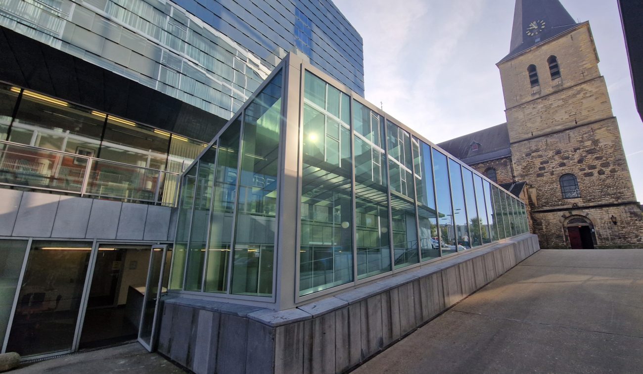 Exterieur SCHUNCK Heerlen – glazen corridor/uitbouw met stalen kozijnen naast de historische kerk met toren, gezien vanaf het pleintje.