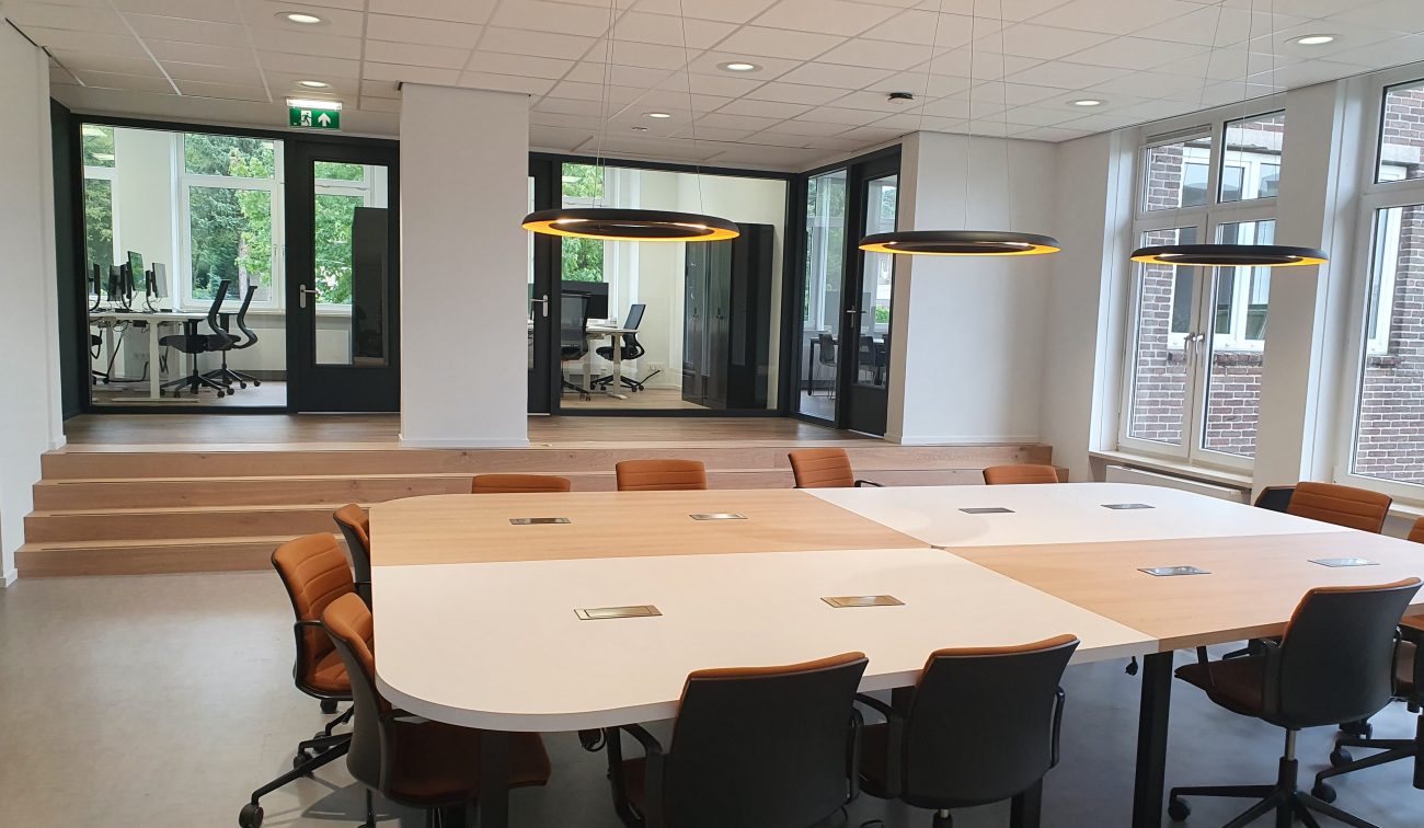 Moderne kantoorruimte met grote vergadertafels en cognackleurige stoelen; drie ronde hanglampen boven de tafels, een verhoogde zone met glazen wanden en deuren op de achtergrond, en hoge ramen die veel daglicht binnenlaten.