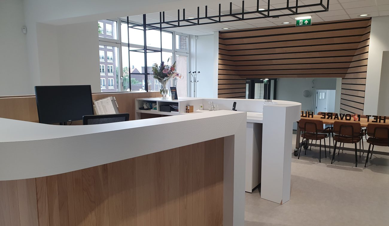 Ontvangstbalie met gebogen wit-houten front in een lichte kantoorruimte; groot raam met daglicht, zwart plafondrooster, houten lattenwand met doorgang en daarachter een vergadertafel met stoelen en glazen deur.