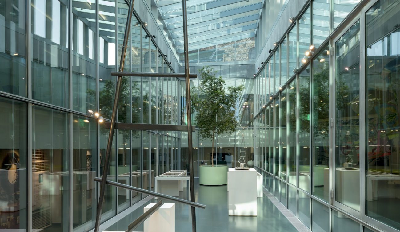 Glazen atrium van SCHUNCK Heerlen met transparant dak; centrale stalen kunstinstallatie op witte sokkel, vitrines en bomen in ronde bakken langs de glazen wanden.