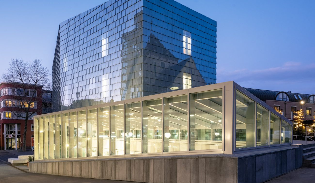 Glazen atrium van SCHUNCK Heerlen met hoge lichtstraat, binnentuin met grote boom en witte vitrines; transparante passerelle en moderne, lichte architectuur.